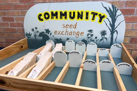 A wooden display holding small packets of seeds with a sign reading Community Seed exchange painted on wood