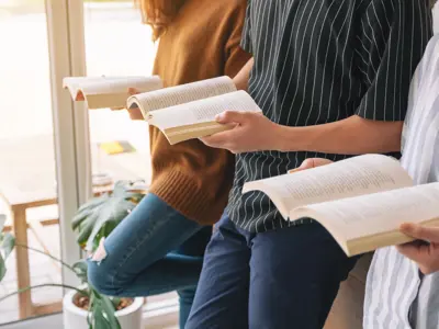 Three people standing by a window reading books